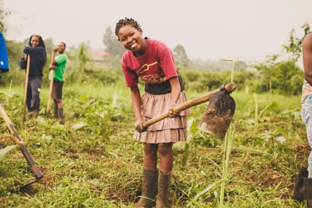 Kira Farm student Jieva smiles at the camera whilst digging the farm land with a hoe